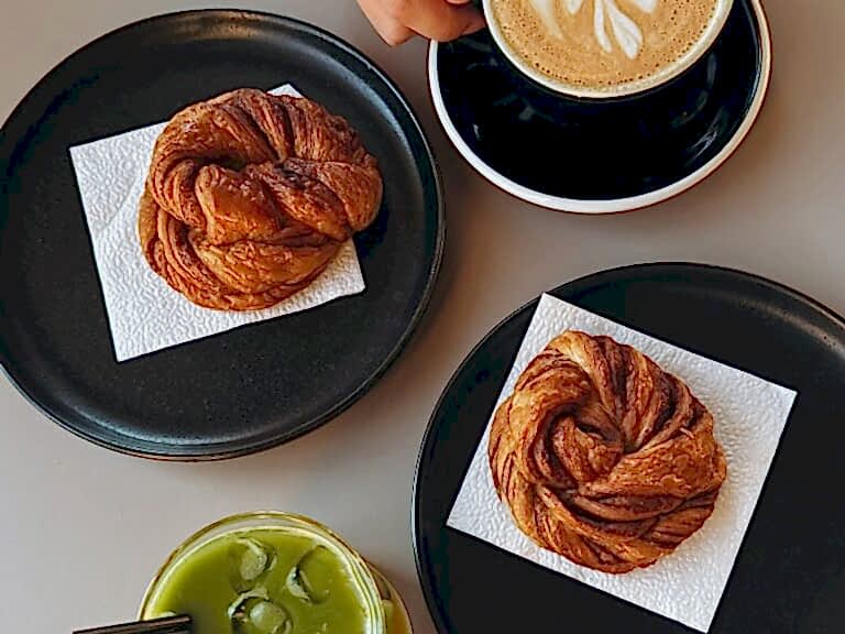 two vegan cinnamon rolls on black plates with white napkins next to a green iced match and a latte at a bakery in berlin