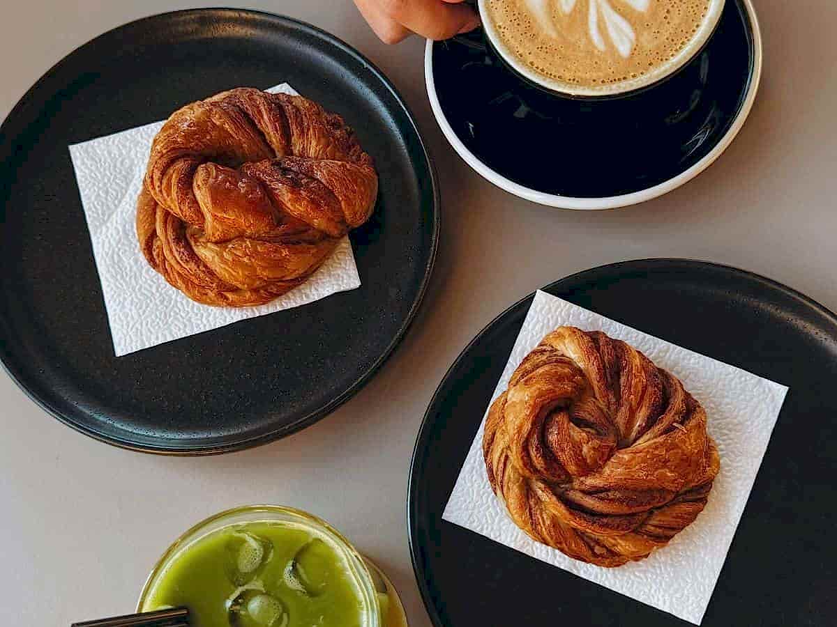 two vegan cinnamon rolls on black plates with white napkins next to a green iced match and a latte at a bakery in berlin