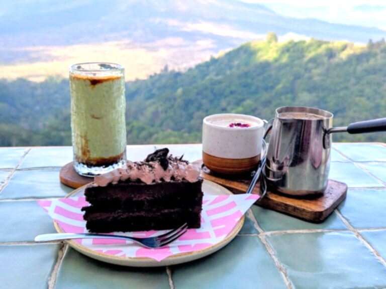 a single slice of dark chocolate vegan cake on a pink plate next to a green avocado iced coffee and a chai tea