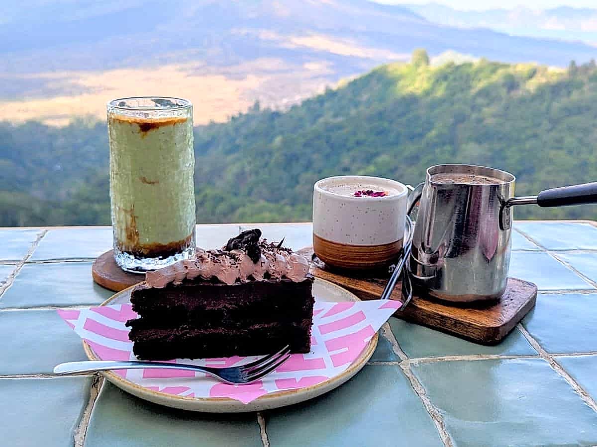 a single slice of dark chocolate vegan cake on a pink plate next to a green avocado iced coffee and a chai tea